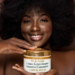 Une personne aux cheveux afro bouclés sourit en tenant un contenant étiqueté « Soin du Corps Or & Argan Crème Éclaircissante Intensive Concentrée Capital Éclat. Dakar