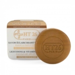 Un pain rond de savon éclaircissant et vitaminé HT26 Paris est placé devant son emballage carré. L'emballage beige et blanc porte le nom de la marque et des informations en français et en anglais. Le savon est de couleur marron clair. Dakar