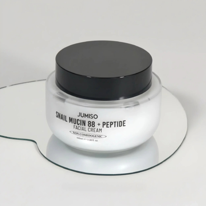 Un pot en verre de crème visage Jumiso à la mucine d'escargot 88 + peptides, doté d'un couvercle noir et d'une étiquette blanche sur une surface réfléchissante. L'étiquette indique qu'elle est non comédogène et pèse 100 ml. Dakar