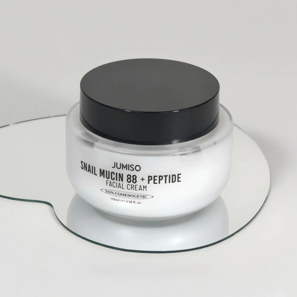 Un pot en verre de crème visage Jumiso à la mucine d'escargot 88 + peptides, doté d'un couvercle noir et d'une étiquette blanche sur une surface réfléchissante. L'étiquette indique qu'elle est non comédogène et pèse 100 ml. Dakar