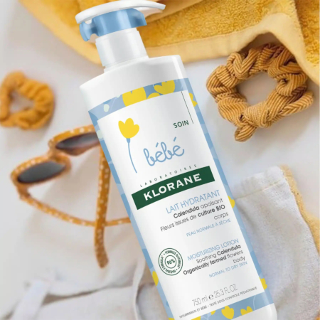 Flacon de lotion hydratante Klorane Bébé avec pompe, orné d'une étiquette bleue et jaune à motifs floraux. Il est posé sur une surface claire, à côté d'une serviette pour bébé, d'une brosse en bois et d'articles pour bébé. Dakar