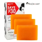 Image d'un trio de savons Kojie San. L'emballage affiche « ÉCONOMISEZ JUSQU'À 30 P » et un visage stylisé. Trois savons orange sont présentés devant la boîte, chacun imprimé du logo Kojie San. Dakar