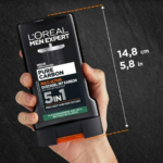 Main tenant un flacon noir de gel douche multi-action Pure Carbon de L'Oréal Men Expert. Le flacon porte une étiquette détaillant le produit. Les dimensions indiquées sont de 14,8 cm x 5,8 pouces. Le fond est une surface sombre texturée. Dakar