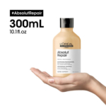 Une main tient un flacon beige de shampoing Absolut Repair de L'Oréal Paris sur fond blanc. Le texte indique « #AbsolutRepair, 300 ml, 10,1 fl. oz ». Le flacon est étiqueté pour un usage professionnel et pour cheveux abîmés. Dakar