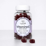 Une bouteille transparente étiquetée « Lashilé Beauty Vitamine E Mono Boost » contient des vitamines rouges en gélifiés. L'étiquette indique « Délicieux gélifiés » et « Vegan | 60 pièces ». Deux gélifiés sont placés à côté de la bouteille sur fond blanc. Dakar