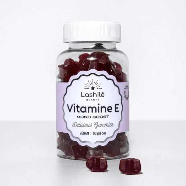 Une bouteille transparente étiquetée « Lashilé Beauty Vitamine E Mono Boost » contient des vitamines rouges en gélifiés. L'étiquette indique « Délicieux gélifiés » et « Vegan | 60 pièces ». Deux gélifiés sont placés à côté de la bouteille sur fond blanc. Dakar