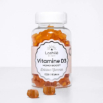 Une bouteille en plastique transparent étiquetée « Lashilé Beauty Vitamine D3 Mono Boost » contient des compléments alimentaires gélifiés orange. L'étiquette mentionne « Délicieux gélifiés », « Vegan » et « 60 pièces ». Quelques gélifiés sont dispersés devant la bouteille. Dakar