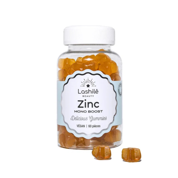 Un flacon transparent étiqueté « Lashilé Beauty Zinc Mono Boost » contient des compléments alimentaires gélifiés orange. L'étiquette indique également « Délicieux Gummies » et « Vegan », avec une mention indiquant 60 gélifiés à l'intérieur. Quelques gélifiés sont placés devant le flacon. Dakar
