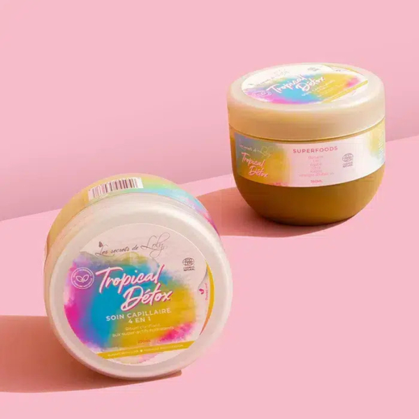 Deux pots de soins capillaires « Tropical Detox » aux couleurs pastel sur fond rose. L'un est vertical, l'autre incliné sur le côté, affichant des étiquettes et du texte colorés. Les ombres projetées par les pots sont visibles en arrière-plan. Dakar