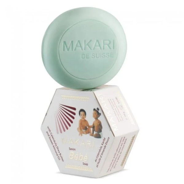 Un savon rond vert menthe, étiqueté « Makari de Suisse », est posé en équilibre sur une boîte hexagonale. La boîte présente une image de deux bébés et un texte sur le savon, intitulé « Savon Bébé ». Dakar