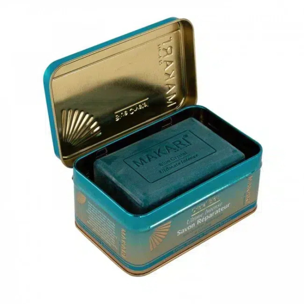 Un pain de savon foncé, étiqueté « Makari », est placé dans une boîte métallique rectangulaire ouverte, à l'intérieur doré et à l'extérieur bleu sarcelle. L'extérieur de la boîte est orné d'un texte doré et de motifs décoratifs. Dakar