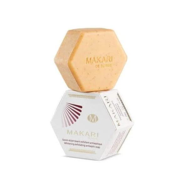 Un pain hexagonal de savon exfoliant antiseptique blanchissant Makari de Suisse est présenté dans sa boîte hexagonale blanche et dorée assortie. La boîte comporte le nom de la marque, le logo et la description du produit en anglais et en français. Dakar