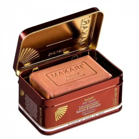 Un pain de savon exfoliant rectangulaire, étiqueté « Makari Exclusif », est présenté dans une boîte métallique à charnière aux accents marron et doré. Le texte sur la boîte souligne ses propriétés exfoliantes et éclaircissantes. Dakar