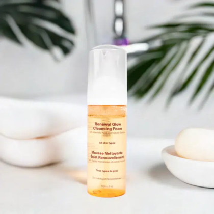 Une bouteille en plastique de « Mousse Nettoyante Éclat Renouveau » au liquide orange est posée sur un plan de travail blanc. En arrière-plan, on distingue un robinet flou et un savon sur une assiette. Dakar