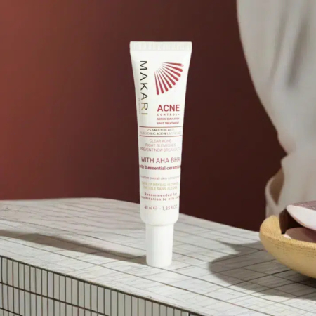 Un tube de crème Makari Acne Control est posé sur une surface quadrillée. Le tube est principalement blanc avec un texte rouge faisant la promotion des AHA et BHA pour le traitement de l'acné. Un arrière-plan flou laisse apparaître une partie d'un bol et un tissu rose. Dakar