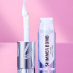 Un tube de gloss transparent et scintillant, doté d'un applicateur blanc brillant, est ouvert sur un fond rose. L'inscription « SHIMMER BOMB » est inscrite en gras. L'emballage arbore un design métallique élégant. Dakar