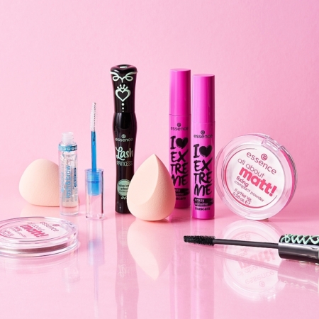 Une collection de produits de maquillage Essence disposés sur une surface rose. Elle comprend des mascaras, un correcteur de couleur, deux éponges à maquillage et des poudres compactes. L'emballage est coloré avec des touches de noir, de rose et de bleu. Dakar