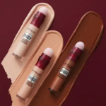 Trois anti-cernes Maybelline Instant Age Rewind sont appliqués sur des échantillons de leurs teintes respectives : claire, moyenne et foncée. Les tubes sont dotés d'applicateurs éponge intégrés et se détachent sur un fond rouge profond. Dakar