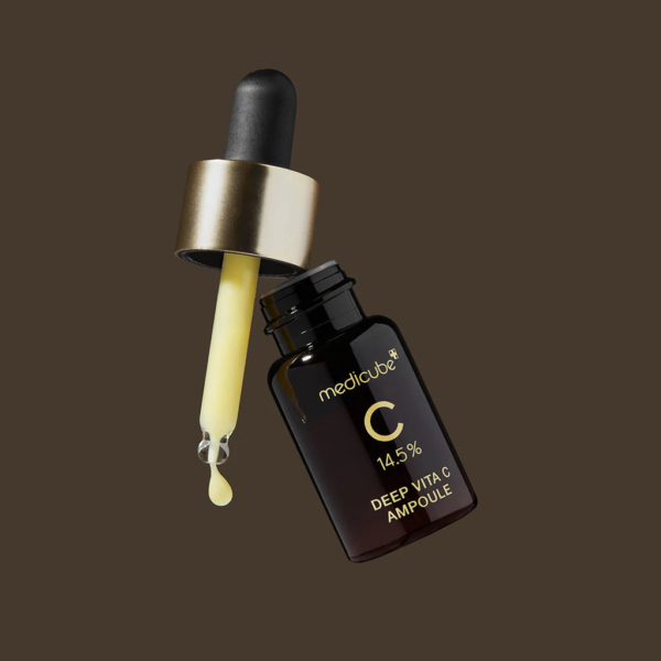 Un flacon compte-gouttes marron portant l'inscription « Medicube Deep Vita C Ampoule 14,5 % » est incliné sur le côté, le compte-gouttes déversant une goutte de liquide jaune. Le flacon et le compte-gouttes se détachent sur un fond marron uni. Dakar