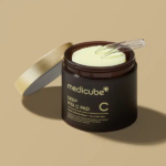 Pot marron foncé de tampons Medicube Deep Vita C, avec couvercle doré légèrement ouvert. À l'intérieur se trouvent des tampons circulaires vert clair surmontés d'un applicateur transparent. Le tout sur fond beige uni. Dakar