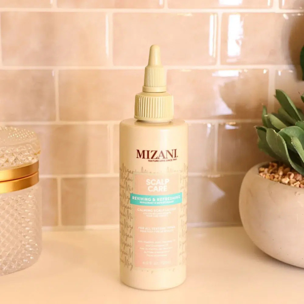 Un flacon de spray revitalisant et rafraîchissant Mizani Scalp Care est posé sur un comptoir de salle de bain avec une crédence en carrelage beige. Le flacon, doté d'un embout pointu, est posé à côté d'une plante décorative et d'un contenant texturé. Dakar