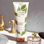 Un tube de gel Mizani True Textures Perfect Coil Oil est posé sur un comptoir de salle de bain, avec un robinet doré et une bouteille en verre décorative en arrière-plan. L'étiquette du produit présente des feuilles vertes et un texte précisant son utilisation pour les boucles, les ondulations et les frisures. Dakar