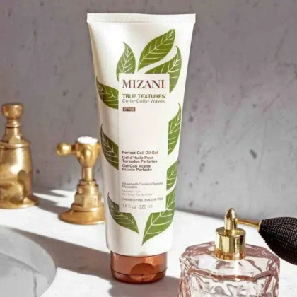 Un tube de gel Mizani True Textures Perfect Coil Oil est posé sur un comptoir de salle de bain, avec un robinet doré et une bouteille en verre décorative en arrière-plan. L'étiquette du produit présente des feuilles vertes et un texte précisant son utilisation pour les boucles, les ondulations et les frisures. Dakar