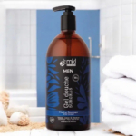 Un flacon marron de gel douche MKL Green Nature, étiqueté « Gel Douche Surgras Men », est posé sur le comptoir de la salle de bain. Il est doté d'une pompe noire et de motifs de feuilles bleues. À l'arrière-plan, des serviettes blanches sont soigneusement empilées. Dakar