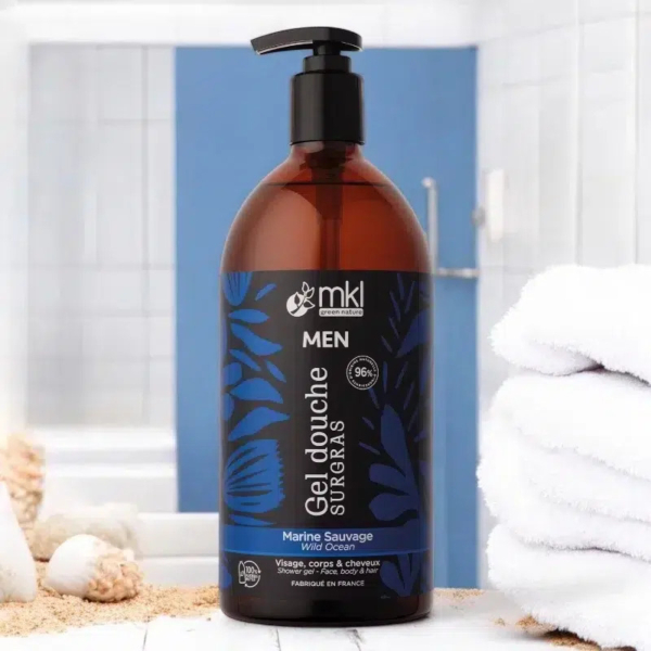 Un flacon marron de gel douche MKL Green Nature, étiqueté « Gel Douche Surgras Men », est posé sur le comptoir de la salle de bain. Il est doté d'une pompe noire et de motifs de feuilles bleues. À l'arrière-plan, des serviettes blanches sont soigneusement empilées. Dakar