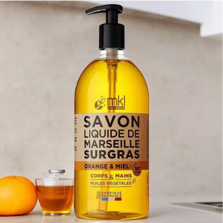 Un flacon pompe de savon liquide de Marseille parfumé à l'orange et au miel est posé sur un plan de travail. L'étiquette comporte un texte en français, avec des images d'une orange et d'une louche de miel. Une orange entière et un petit pot de miel apparaissent en arrière-plan. Dakar