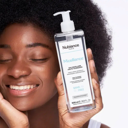 Une personne aux cheveux bouclés sourit, les yeux fermés, tenant un flacon transparent d'eau micellaire Nubiance Paris Micelliance. Le flacon est muni d'une pompe et est étiqueté pour un nettoyage en douceur. La peau est lisse et éclatante. Dakar