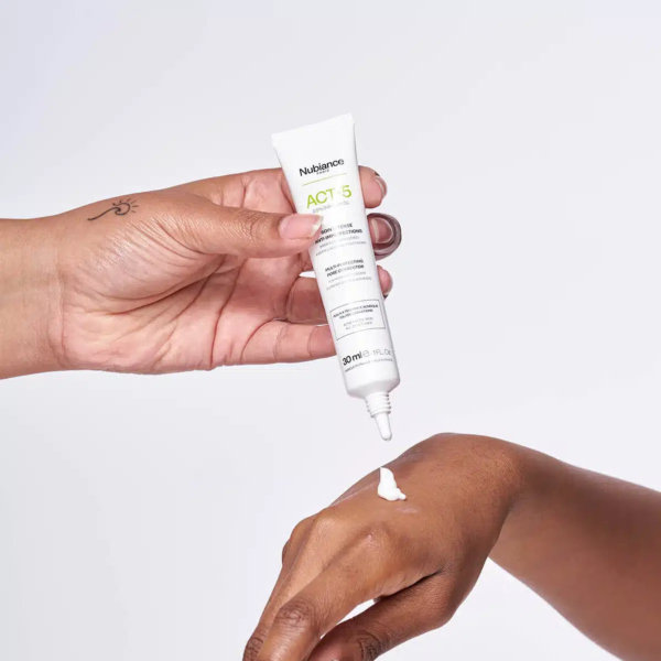 Une personne applique une petite quantité de crème blanche provenant d'un tube étiqueté « ACT 5 » sur le dos de la main d'une autre personne sur un fond blanc uni. Dakar