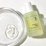 Un flacon pompe étiqueté « numbuz:n serum 5 - Goodbye Blemish » est posé à côté d'un plateau en verre circulaire ouvert contenant un tourbillon de crème blanche. Le flacon et le plateau reposent sur une surface blanche. Dakar
