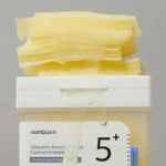 Un contenant de soin pour la peau étiqueté « Vitamine-Niacinamide Concentré » avec une épaisse substance gélatineuse jaune qui déborde. L'étiquette indique « Antioxydant Éclaircissant 5+ ». Le fond est d'une couleur unie et neutre. Dakar