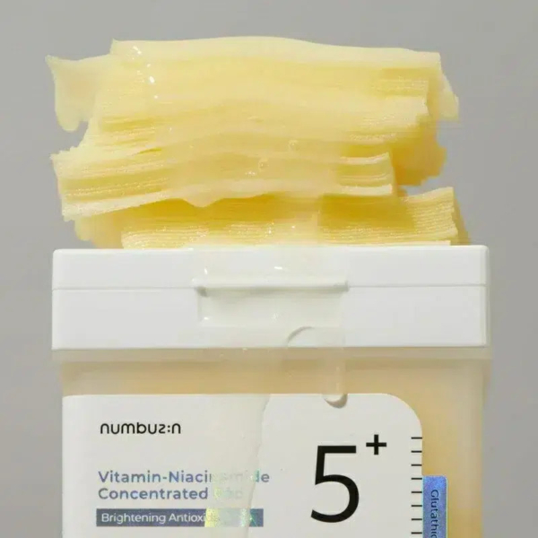 Un contenant de soin pour la peau étiqueté « Vitamine-Niacinamide Concentré » avec une épaisse substance gélatineuse jaune qui déborde. L'étiquette indique « Antioxydant Éclaircissant 5+ ». Le fond est d'une couleur unie et neutre. Dakar