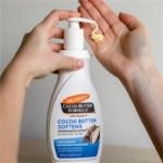 Une main distribue une lotion d'un flacon blanc étiqueté « Palmer's Formule au beurre de cacao avec vitamine E », conçue pour adoucir et soulager la peau sèche. Le flacon est tenu et une petite quantité de lotion est déposée sur les doigts de la main opposée. Dakar