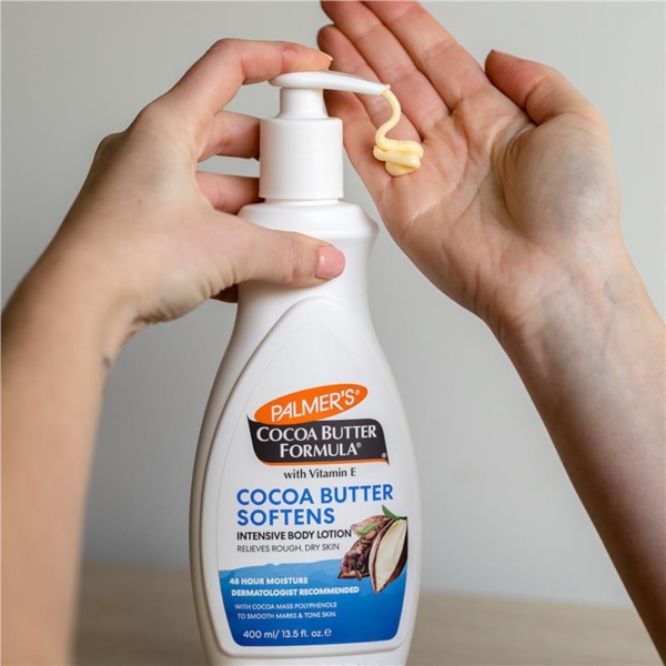Une main distribue une lotion d'un flacon blanc étiqueté « Palmer's Formule au beurre de cacao avec vitamine E », conçue pour adoucir et soulager la peau sèche. Le flacon est tenu et une petite quantité de lotion est déposée sur les doigts de la main opposée. Dakar