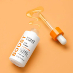 Un flacon de Super Booster C15 de Paula's Choice repose sur une surface orange. Le flacon est blanc avec un texte orange, et à côté se trouve un compte-gouttes orange contenant du sérum déversé en zigzag. Dakar