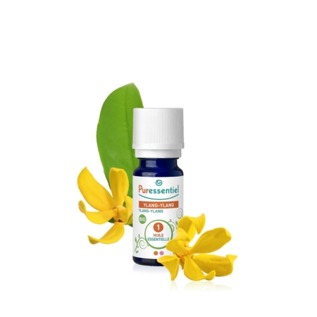 Un flacon d'huile essentielle d'Ylang-Ylang Puressentiel est présenté, avec un bouchon blanc et une étiquette verte. Deux fleurs d'Ylang-Ylang jaunes et une feuille verte sont disposées autour du flacon sur fond blanc. Dakar
