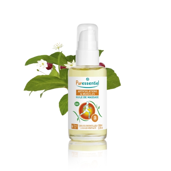 Un flacon d'huile de massage Puressentiel pour les articulations et les muscles, contenant un liquide transparent. L'étiquette est blanche et orange. Derrière le flacon, un arrangement de feuilles vertes et de petites fleurs blanches. Dakar