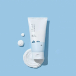 Un tube de crème de soin au design blanc et bleu clair est posé sur un fond bleu clair. Le produit est accompagné de textures onctueuses, une en tourbillon et deux en petites doses. Dakar