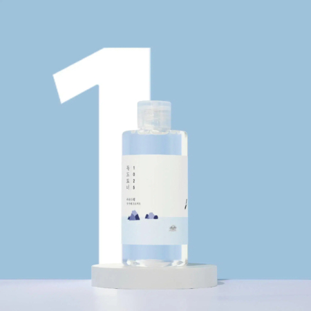 Une bouteille transparente, ornée d'une étiquette minimaliste ornée de petits motifs de montagnes, est placée sur un fond bleu clair. Le chiffre « 1 » est bien visible en arrière-plan, la bouteille reposant sur une plateforme circulaire blanche. Dakar