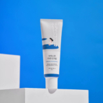 Un tube blanc de crème de soin, orné d'un texte coréen et d'une illustration minimaliste, est posé sur une plateforme blanche. Le fond bleu ciel uni crée une esthétique épurée et fraîche. Dakar