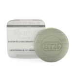 Un pain de savon rond vert avec l'inscription « HT26 PARIS » en relief se trouve à côté de son emballage blanc avec un texte bilingue, étiqueté « HT26 Savon Éclaircissant Caviar », et offre des bienfaits éclaircissants et riches en vitamines pour la peau. Dakar