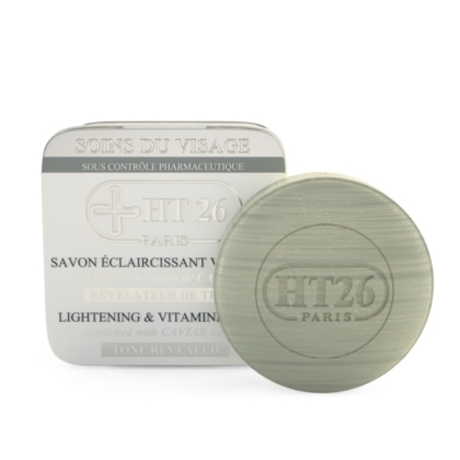 Un pain de savon rond vert avec l'inscription « HT26 PARIS » en relief se trouve à côté de son emballage blanc avec un texte bilingue, étiqueté « HT26 Savon Éclaircissant Caviar », et offre des bienfaits éclaircissants et riches en vitamines pour la peau. Dakar