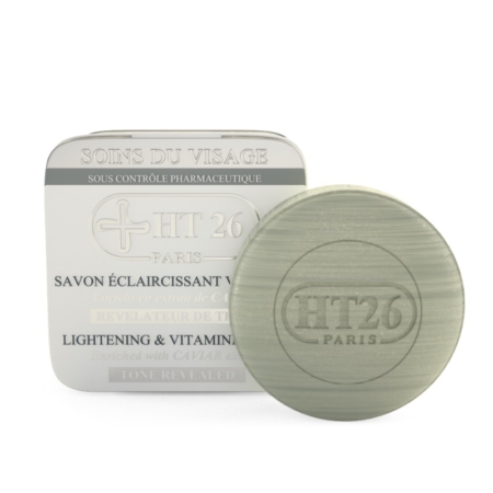 Un pain de savon rond vert avec l'inscription « HT26 PARIS » en relief se trouve à côté de son emballage blanc avec un texte bilingue, étiqueté « HT26 Savon Éclaircissant Caviar », et offre des bienfaits éclaircissants et riches en vitamines pour la peau. Dakar