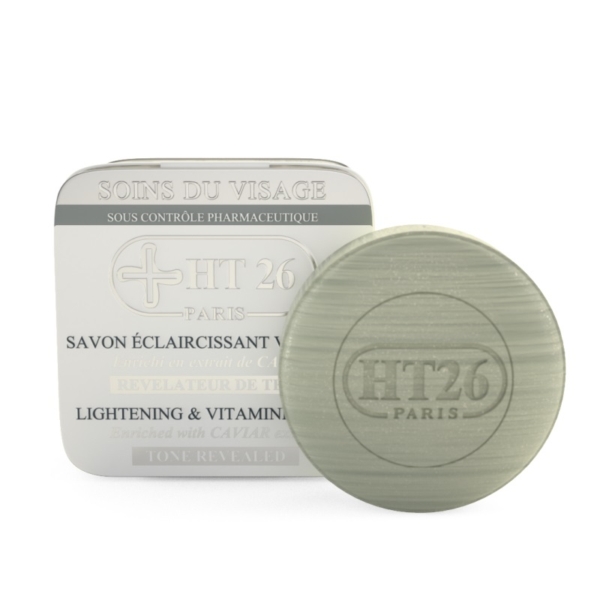 Un pain de savon rond vert avec l'inscription « HT26 PARIS » en relief se trouve à côté de son emballage blanc avec un texte bilingue, étiqueté « HT26 Savon Éclaircissant Caviar », et offre des bienfaits éclaircissants et riches en vitamines pour la peau. Dakar