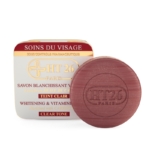 Un savon rond marron estampé « HT26 Paris » se trouve devant une boîte carrée blanche et dorée étiquetée « HT26 Savon Blanchissant Carotte » avec un texte rouge et un symbole de croix, soulignant sa promesse Blanchissant & Vitamine C. Dakar