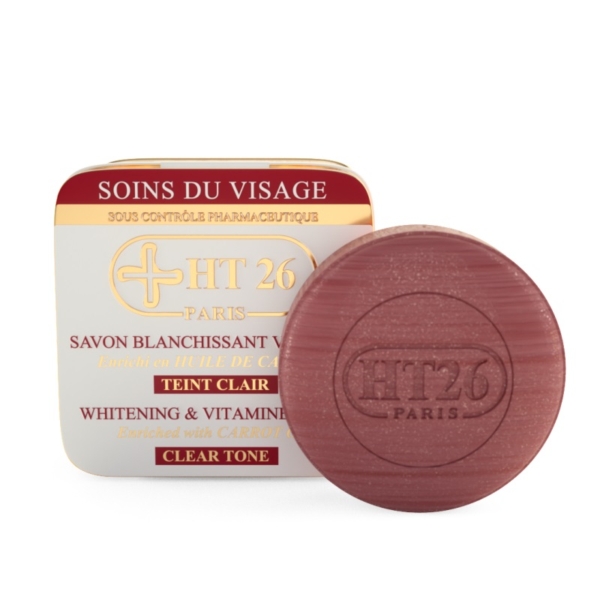 Un savon rond marron estampé « HT26 Paris » se trouve devant une boîte carrée blanche et dorée étiquetée « HT26 Savon Blanchissant Carotte » avec un texte rouge et un symbole de croix, soulignant sa promesse Blanchissant & Vitamine C. Dakar