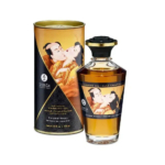 Un flacon d'huile chauffante Shunga Erotic Art est présenté à côté de son emballage cylindrique. Tous deux sont ornés d'une illustration traditionnelle japonaise représentant un couple enlacé. L'étiquette porte l'inscription « Baisers Caramel » et l'huile est de couleur ambrée. Dakar